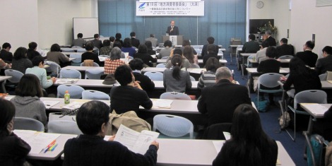 着席している参加者の後ろから会場全体を撮影した写真。前方の演台には河上委員長がいる