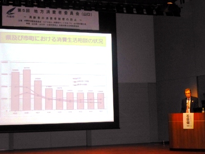 ステージ上には「県及び市町における消費生活相談の状況」というグラフが映し出された大きなプロジェクタ、その横で石田所長が報告をしている写真